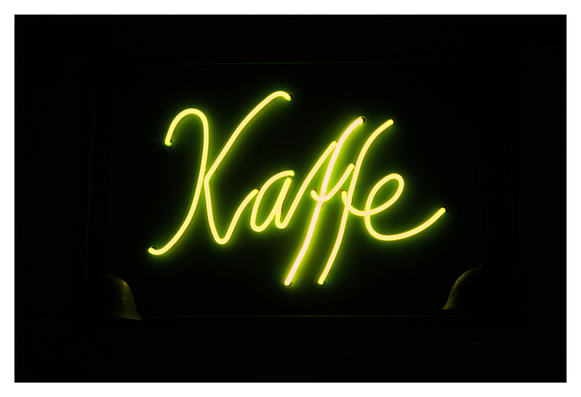 En liggande bild på en gul neonskylt med texten ”Kaffe” mot en svart bakgrund. I de nedre hörnen av skylten faller lite ljus på två armé-hjälmar.