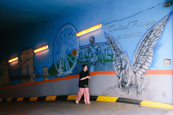 A person walks in front of a mural, containing a horse with wings, a soccer player and some landmarks. A text states Central Passage Budapest.