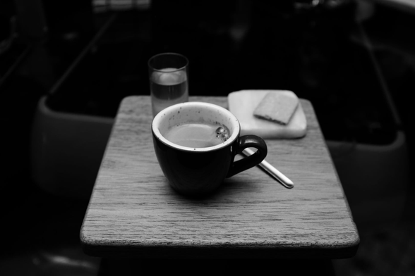 Fotografía en blanco y negro que muestra una taza de café oscuro sobre una pequeña mesa de madera. A su lado, una cucharilla metálica, un vaso de agua y una galleta sobre una servilleta. Los tonos predominantes son oscuros, con contraste marcado entre sombras profundas y superficies claras.

Black and white photograph showing a dark coffee cup on a small wooden table. Beside it, a metal spoon, a glass of water, and a cookie on a napkin. The predominant tones are dark, with strong contrast between deep shadows and lighter surfaces.