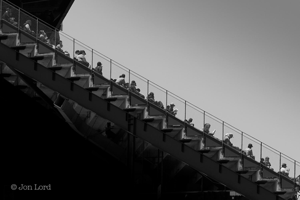 This is a black and white photo in landscape format of the tiered stands (bleachers) in a sports stadium viewed edge on. Oracle Park, San Francisco (2015).
The image is divided in half diagonally. The lower part forms a dark triangle radiating from the lower left corner upwards and to the right. Stretching across the frame from the upper left corner down to the lower right corner is the tiered sports spectator stand, dividing the image in half. In view is a dark, stout metal girder supporting the seating area. Above the girder is a series of zig-zag, tiered  steps where the seats are located. Above this is a short fence of a little over a metre. Behind the fence are a small number of spectators, a few on each tier. In the upper right corner is a darkened, clear and cloudless sky. The game is yet to start.