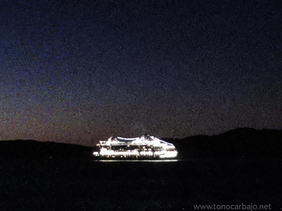 Foto nocturna de un transatlántico saliendo de la Ria de Vigo. El transatlántico esta iluminado, un poco fuera de foco, es casi una mancha blanca irreal en el oscuro de la noche. El mar y la costa están en negro el cielo conserva aún un poco de luz del atardecer.
La foto tiene grano visible, se ve la textura lo que le da un aspecto pictórico
