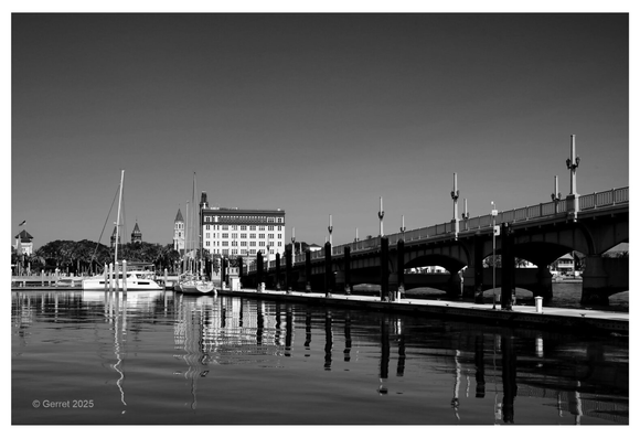 Black and white image of a waterfront with sailboats docked alongside a calm body of water. A bridge on the right leads towards buildings, evoking tranquility.