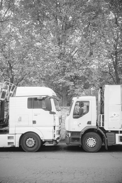 Black and white portrait format photo of the white cabins of 2 parked trucks facing each others. Leaves of trees are behind