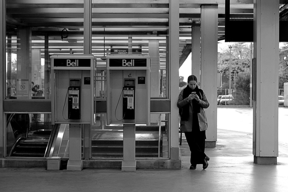 On the outdoor platform for buses and streetcars, a woman stands in front of the structure covering the way down to the subway platforms. She is focused intently on her phone, standing beside two unused Bell payphones.