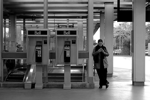 On the outdoor platform for buses and streetcars, a woman stands in front of the structure covering the way down to the subway platforms. She is focused intently on her phone, standing beside two unused Bell payphones.
