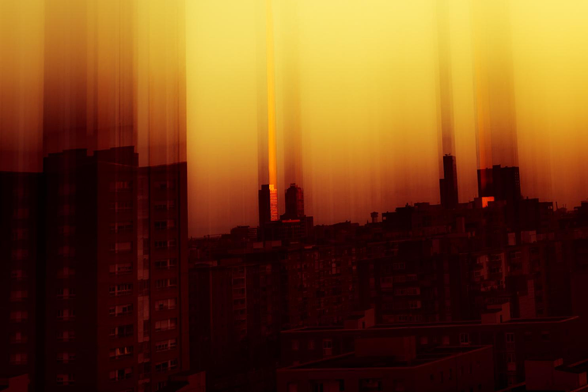Foto de un atardecer urbano en Barcelona. Los rayos de sol dorados, se filtran entre los edificios altos, creando un efecto de luz difusa y vertical que ilumina el cielo con tonos anaranjados y ocres. En el medio se adivinan las torres Mapfre. En primer plano un edificio de viviendas del barrio. 
Las siluetas oscuras de los edificios se difuminan hacia arriba y contrastan fuertemente con la luminosidad del cielo, dando un dramatismo teatral a la imagen.