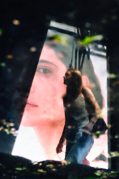 A person is seen through a reflection of a puddle scattered with green leaves blurry in the foreground. The person has long blonde hair wearing a tank top and jeans. They are walking in front of a large poster of a feminine face that is half illuminated and half in darkness.