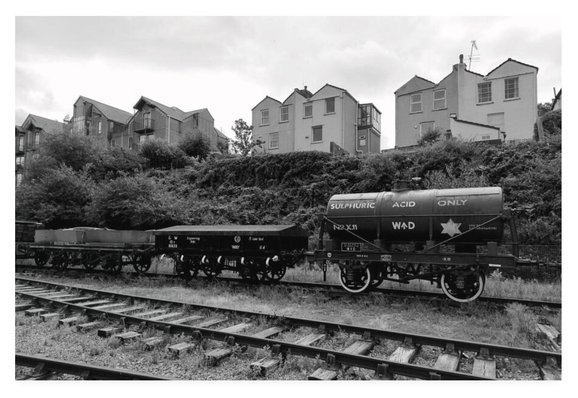 Black and white photograph of railway tracks with old rolling stock.  There is a tanker marked Sulphuric Acid Only attached to flatcars.  Above there are regular domestic homes.