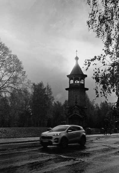 The photo shows a church with a bell tower surrounded by trees. A car is driving on the road in front of the church. The image is in black and white. The dome is illuminated by the sun behind it.
