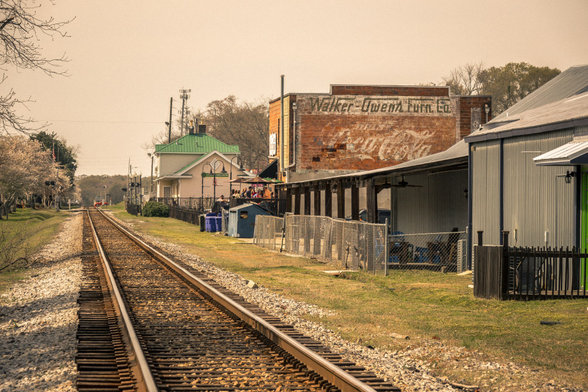 Railroad tracks running through a small town with historic buildings on the right side. In the distance, a cream-colored building with a green metal roof and arched windows sits near the tracks. On the right, weathered corrugated metal buildings with green trim line the railway, behind which stands a two-story red brick building with faded vintage painted advertising text reading "Walker-Owens Furn Co." and additional text below. The scene includes bare trees, utility poles, a cell tower in the distance, and chain-link fencing separating the tracks from the buildings. The lighting suggests late afternoon with a hazy, warm-toned sky.
