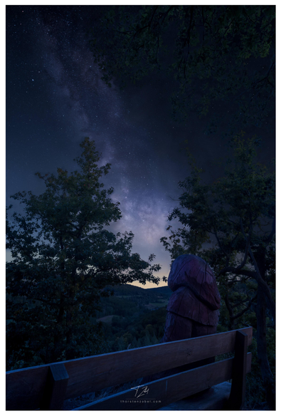 A wooden man wearing a hood sits on a bench at night, looking out over a valley. Above the valley, the Milky Way is clearly visible, framed by oak trees in the foreground.