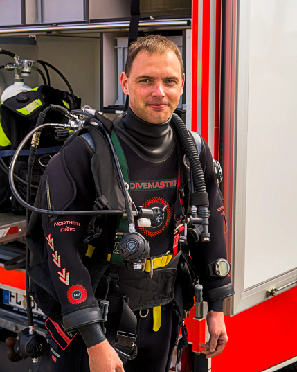 Ein Taucher der Berufsfeuerwehr Flensburg steht in voller Montur vor einem Ausrüstungswagen der Feuerwehr und lächelt in die Kamera. 
. 
A diver from the Flensburg professional fire brigade stands in full gear in front of a fire brigade equipment truck and smiles at the camera. 
. 
