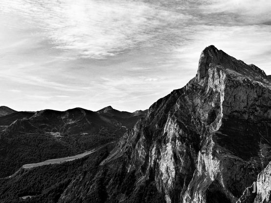 Fotografía en blanco y negro de un paisaje montañoso. Un imponente pico rocoso y escarpado domina el lado derecho, mostrando paredes verticales con texturas rugosas y marcadas por la luz y la sombra. Hacia el centro y la izquierda, una vasta extensión de bosques densos cubre las laderas de montañas más bajas que se extienden hacia el horizonte. Una franja clara y alargada, posiblemente un prado o un sendero, atraviesa el denso follaje en el valle. El cielo ocupa la parte superior, con nubes finas y alargadas que añaden textura y profundidad. La ausencia de color realza las formas, las texturas y el contraste dramático entre la roca, la vegetación y el cielo.