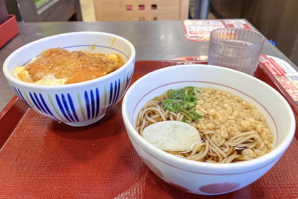 Una bandeja sobre una mesa de un local de una cadena de comida rápida japonesa (Nakau) y sobre ella un bol de fideos de soba, otro bol con un katsudon (lomo empanado y frito cortado en tiras sobre arroz blanco y cubierto de huevo y cebolla) y un vaso de plástico con agua.