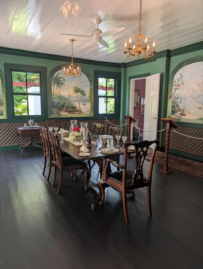 We're in one corner of a dining room in a colonial style house. The floors are dark wood, smooth enough to reflect light coming in from the windows on one wall opposite us. The walls are painted light green while the window frames stand out with a darker shade. Between the two windows and on the adjacent wall too there are painted murals with arched tops, both depicting tropical island scenes. A rectangular dining table in dark wood seats six and the table is set for a formal dinner. Through the windows opposite we can see bright sunlight hitting lush foliage.