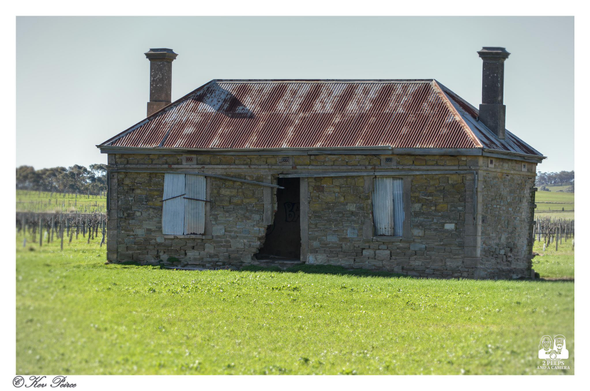 Stone ruins of an old, abandoned farmhouse with a rusted corrugated iron roof, two tall brick chimneys, and boarded up windows, set in a vibrant green field.

The stone walls are partially crumbled around the front doorway, revealing the darkness inside. A vineyard and trees are visible in the background under a pale blue sky. Signed Kev Peirce.