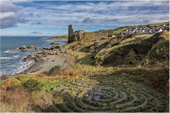 Scenic coastal view featuring a rocky shoreline, ruins of a castle on a grassy hillside, and a small village in the background under a partly cloudy sky. A circular stone labyrinth is visible in the foreground.