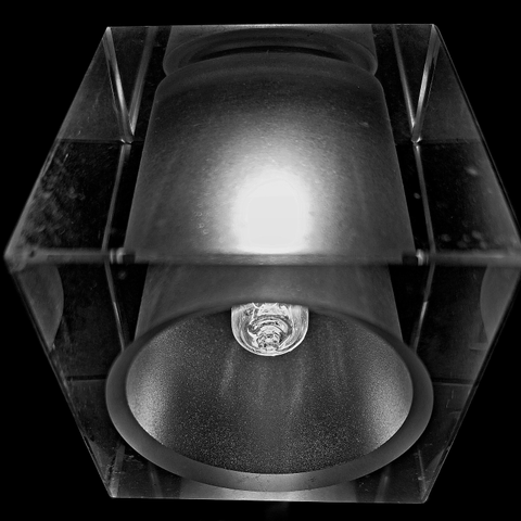 Macro capture from a lower angle: A square lamp made of clear glass. In the centre of the lamp is a frosted glass cylinder containing the light bulb. The light from the bulb is reflected by the cylinder as a luminous oval.  Around the lamp, it is pitch black night. The hard, clear shape of the luminaire conveys coldness. Only the glow of the light breaks through this austerity.