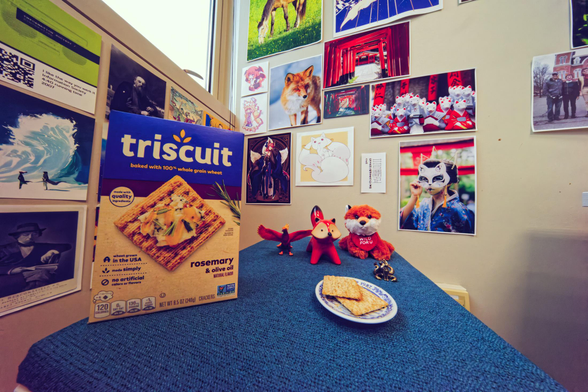 At the bottom a blue  rectangular surface is made to look like a trapezoid thanks to wide angle distortion, to left of it box of triscuits is cocked at a funny angle and to the right there are two on a little plate and three fox figurines and a phoenix.  Behind it to the left side of a corner are prints including an information society album cover, kung fu people in front of a wave and Benjamin Disraeli,  to the right cartoon pictures of fictrional fox spirts, a real fox, and scenes at an inari shrine in Japan