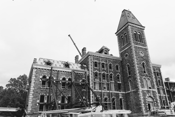 Black and wehite photo of an old stone building trimmed lighter on the corners than the rest with a huger steel framework entering it from the front and a crane visible on the other side.