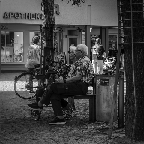 Man sieht eine Szenen in einer Fußgängerzone. Ein Renterehepaar sitzt auf einer Bank und erholt sich, während um sie herum die Menschen weiter gehetzt an ihnen vorbeigehen. Das Foto ist in Schwarzweiß.