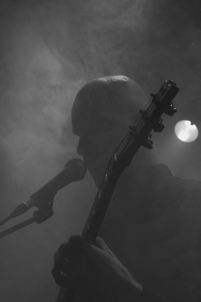 Ein Mann mit E Gitarre steht vor einem Mikrofon und singt hinein. Im Hintergrund erkennt eins ein Scheinwerfer der durch den Bühnenennebel leuchtet 