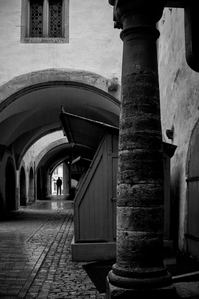 The image is in black and white and shows an arched passageway with cobblestone pavement. In the foreground, there is a large stone column on the right, next to wooden structures with slanted roofs. In the background, there are more round arches and a single person standing in the light, visible as a silhouette. At the top left, a window with small round decorations is visible.