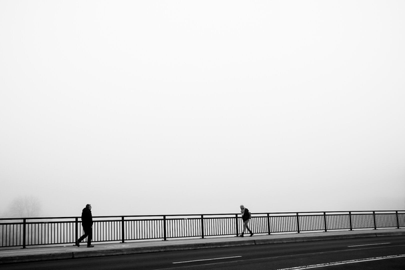 The black-and-white photograph shows a foggy scene with two people walking in opposite directions on a bridge. The bridge has a metal railing, and the road in the foreground appears empty. The background is dominated by thick fog, obscuring any additional details such as landscape or buildings.