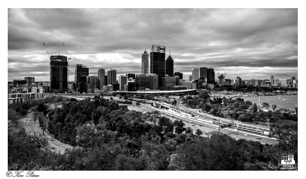 A black and white, panoramic photograph of the Perth, Western Australia central business district skyline, viewed from a high vantage point (Kings Park).

A multi lane freeway (Kwinana/Mitchell Freeways) and rail lines cut through a dense foreground of parkland and trees, leading up to the cluster of modern skyscrapers under a heavily clouded, dramatic sky.

The Swan River is visible on the far right.