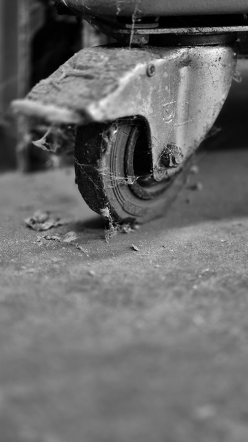 Auf dem Schwarzweißfoto ist eine verschmutzte Lenkrolle mit Bremse von einem Werkstattwagen zu sehen. Die Lenkrolle wurde schräg von der Seite, bodennah, aufgenommen. An ihr haftet Schmutz sowie Spinnweben. Der Boden vor ihr ist ebenfalls verunreinigt.