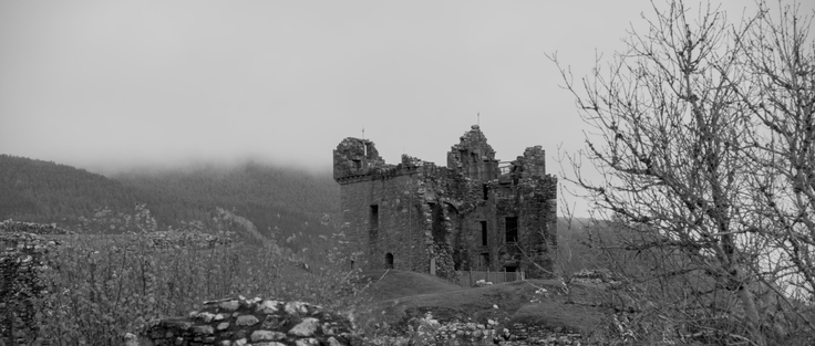 Black and White photo of the ruins of a castle. A great part of the building is missing but some walls are still there. Around it, nature, with the Highlands in the background