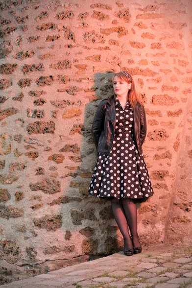 A young woman with red hair, a headband on it, wearing a black dress with white dots reminiscent of the 50s, a black leather jacket and black shoes. She’s leaning against a stone wall on a tilted floor, looking far away, squinting her eyes. An orange light is coming from the top right of the frame
