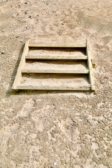 Eine vierstufige Holztreppe liegt mittlerweile flach auf dem Strand im Sand. Der Sand ist durch Wind und Ebbe und Flut so versetzt worden, dass kein Höhenunterschied mehr vorhanden ist und nur noch der vordere Teil der Treppe sichtbar ist. Ansonsten ist sie von Sand bedeckt. 