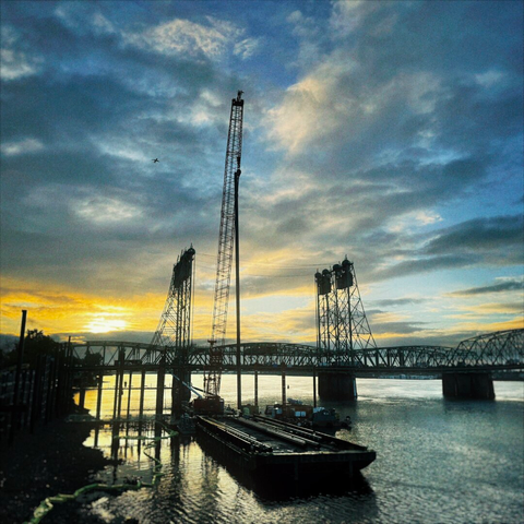 A giant crane assembles foundational pieces of a new structure directly next to the I5 Bridge connecting Oregon and Washington, as an airplane flies overhead against a bright orange and blue sunrise on a partly cloudy morning.