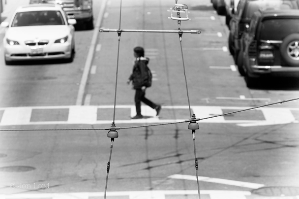 This is an abstract black and white photo of a woman crossing a city street while viewed through the overhead power cables for the municipal Trolleybus. San Francisco (2015).
In the centre of the image is a lady crossing a city street, she is viewed in profile and walking on a pedestrian crossing from right to left. While she is blurred out of focus, she can be seen wearing a black anorak and dark trousers. Along the right margin of the photo is a row of parked cars, on the left margin and the opposite side of the road is a line of cars facing the camera and waiting for the lady to cross or the lights to change. Framing our lady and in focus are two parallel rows of electrical transmission cables stretching from the base of the image, through the centre and up to the top of the frame overhead the street. Below the centre of the photo is a smaller cable stretched at right angles across the street and with two insulators, is supporting the main cables. Above centre is a metal bar that keeps the parallel cables parallel. Its between the bar and the insulators that the lady is framed in the photo.
The location: Stockton Street in San Francisco's Chinatown.