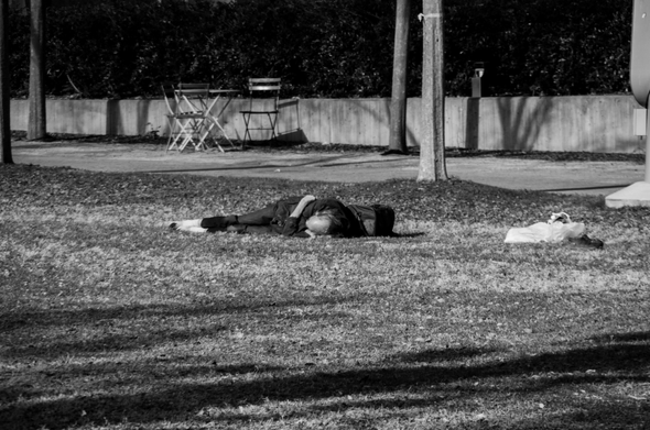 A homeless man sleeping on the ground of a downtown park with all his worldly possessions nearby.