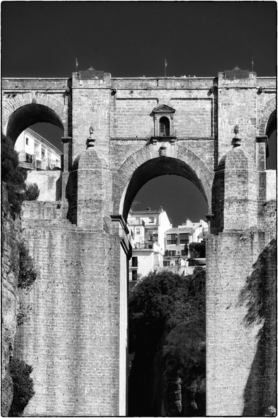 Eine beeindruckende Schwarz-Weiss-Aufnahme des berühmten Aquädukts von Ronda, Spanien. Die massiven Steinbögen des Bauwerks ragen hoch über eine tiefe Schlucht und verbinden die beiden Felswände. Im Vordergrund dominieren drei grosse, runde Bögen, die sich durch ihre klare, geometrische Form abheben. Durch den mittleren Bogen hindurch erkennt man im Hintergrund weisse Häuser, die sich an den Hang schmiegen und von üppiger Vegetation umgeben sind. Die Felswände der Schlucht sind steil und von Pflanzen bewachsen, was einen starken Kontrast zu den glatten Steinbögen bildet. Das Licht fällt von oben ein und betont die Struktur und Tiefe der Architektur.