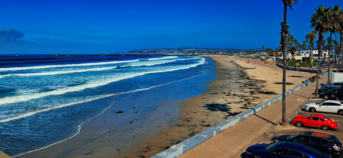 This is a wide, panoramic view of a beach scene on a sunny day. The foreground features a sandy beach with gentle waves rolling in towards the shore. A concrete barrier runs along the edge of the beach, separating the sand from a paved road. Rows of parked cars, including a red convertible and several SUVs, line the road. Palm trees are scattered along the beach and in the distance, and a few people can be seen walking along the shoreline. In the far background, a line of buildings and hills completes the scene.