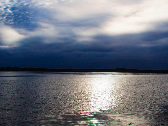 The image depicts a body of water, likely a lake or ocean, under a cloudy sky. The water’s surface reflects the sky, creating a shimmering effect across the frame. Dark, undefined shapes are visible along the horizon line, suggesting land or distant hills. The sky is filled with layers of gray and white clouds, creating a dramatic and somewhat moody atmosphere. The overall tone of the image is subdued and focuses on the contrast between the dark land and the reflective water and sky. 