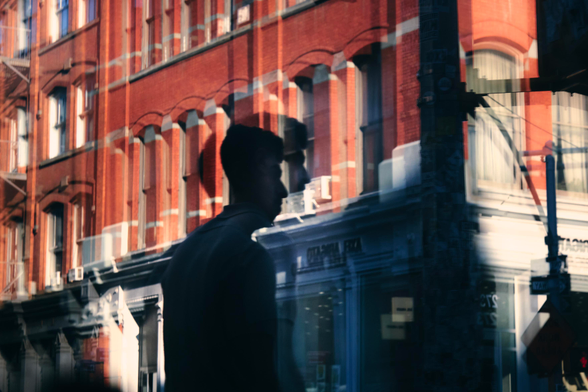 A silhouette is seen through a reflection, repeated and layered as if many shots were taken and blended together. The person stands in front of a red brick building with a white awning for a store below. Next to them is a pole with signage attached.