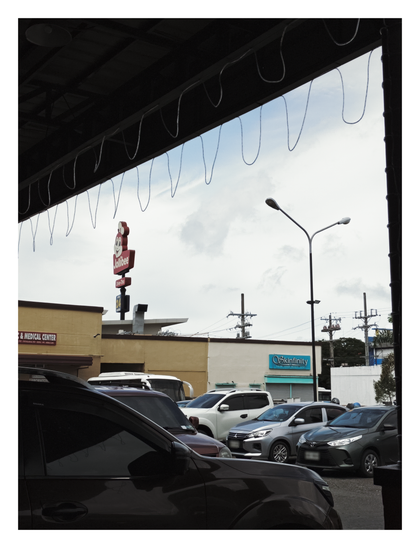 A parking lot with several cars, viewed from under a covered area. In the background, there are a few commercial buildings. A sign for Jollibee is visible in the center, and a sign for "Skinfinity" is on a building to the right of it. The sky is partly cloudy. - Google Lens' AI Overview