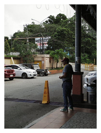 A man in a red baseball cap, grey t-shirt, and blue jeans stands on a tiled sidewalk, looking out onto a parking lot on an overcast day. Several cars, including a red hatchback, a white sedan, and a black jeep, are parked on the asphalt. A yellow caution cone is placed on the ground near the man, and a recycling bin stands next to a pillar. In the background, there are lush green trees and a building with a banner. - Google Gemini 2.5 Pro