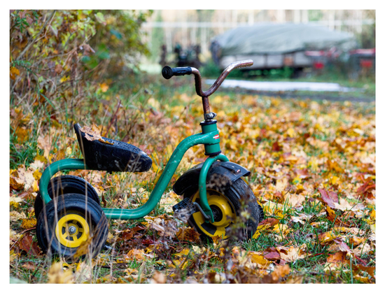 En liggande bild på en grön trehjuling till cykel för små barn. Den står på en gräsmatta täckt med löv i alla höstfärger. I bakgrunden står en båt med kapell på en kärra.