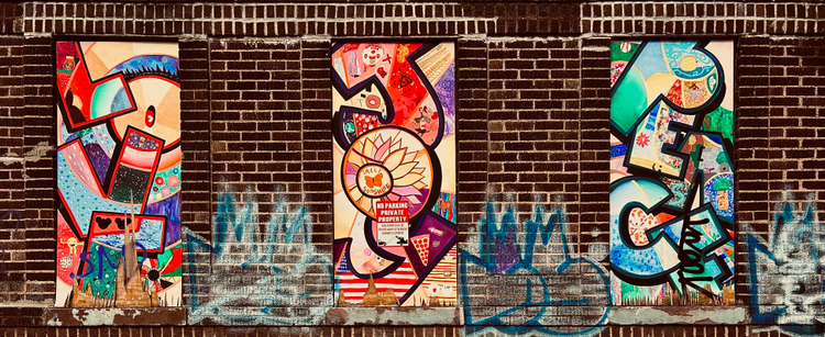A color photograph in landscape orientation of a street art installation in the Bay Ridge neighborhood of Brooklyn. The scene consists of a section of a dark brick wall featuring three rectangular, vertical murals installed within former window openings. Each mural is filled with vibrant, overlapping abstract and illustrative elements in a dynamic, collage-like style. The brickwork is dark brown/red and runs across the top and between the murals, while the bottom section of the wall below the murals shows signs of blue and light-colored graffiti tags that are partially washed out or faded. From left to right the murals spell out the words: love, joy, peace. The leftmost mural primarily uses tones of red, blue, purple, and yellow/tan, featuring bold, curved, and angular shapes. The center mural is dominated by reds, oranges, and pinks, and prominently features a large, stylized sunflower with a butterfly and the text ‘Sally Sunshine’ wrapped around it. A small red and white sign is visible over the flower, stating "NO PARKING PRIVATE PROPERTY. Violators will be towed away at owner’s expense." The rightmost mural is characterized by vivid greens, teals, and blues. All three murals have incorporated within the letter shapes many smaller details such as floral and technological elements. It is day time and sunlight is illuminating the scene, but not light casts or shadows are visible in the scene.