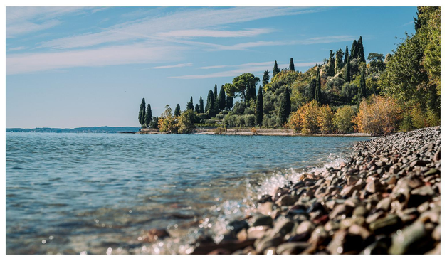 Links ein blauer See, rechts ein Strand mit grauem Kies, in einiger Entfernung ein von Zypressen bewachsener Hain, darüber ein blaudiesiger Himmel. Hach.