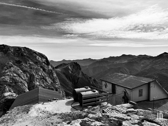 Una imagen en blanco y negro de un vasto paisaje montañoso bajo un cielo con nubes dispersas y una notable estela de condensación de avión. En el fondo, múltiples capas de montañas se extienden hasta el horizonte, volviéndose más pálidas y difusas con la distancia.

El primer plano y el medio muestran un terreno rocoso y árido. A la derecha, se alza un edificio funcional de una sola planta con un techo inclinado y ventanas, que sugiere una estación o refugio de alta montaña. Frente a este, a la izquierda, hay dos grandes tanques cilíndricos sobre una estructura metálica, probablemente para almacenamiento. A la izquierda de los tanques y dominando el lado izquierdo de la imagen, una escarpada y rocosa ladera de montaña se eleva abruptamente. Debajo de esta ladera, se aprecia una segunda estructura más pequeña y angular con un techo oscuro y puntiagudo, parcialmente integrada en el terreno. La escena evoca una sensación de aislamiento y robustez en la alta montaña.