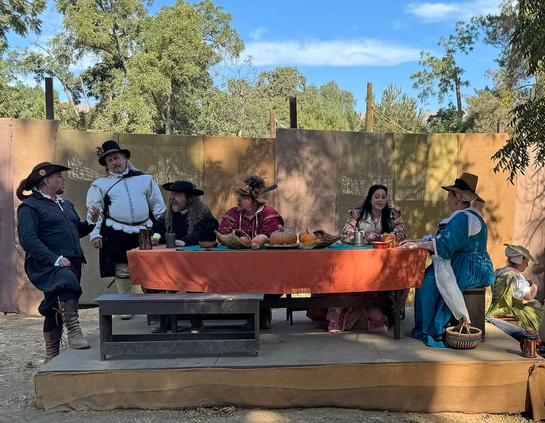 Several people in renaissance period costumes and engaged in conversation sit or stand at a long outdoor eating table with bench seating.