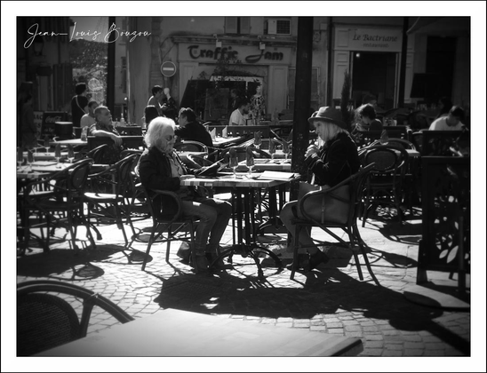 The image captures a serene outdoor café scene bathed in sunlight, creating long, intricate shadows on the cobblestone pavement. Two individuals sit opposite each other at one of the round metal tables, deeply engaged in conversation. The older person on the left, with white hair, reclines comfortably in their chair, batting sunlight off their face, which contrasts strikingly with the darker shadowy tones around them. Opposite, the other person, dressed casually with a hat, leans slightly forward, perhaps listening intently or sharing a story, adding a sense of intimacy to the moment.
The surrounding tables, mostly empty, hint at a quiet moment in what might usually be bustling with patrons. The wrought-iron chairs and tables evoke a classic European café vibe, enhancing the timeless quality of the scene. In the background, blurred storefronts and passersby suggest an urban setting, adding life and context without distracting from the central pair.
The image's black-and-white palette not only heightens the play of light and shadow but also imbues the scene with a nostalgic, almost poetic ambiance. This monochromatic choice invites viewers to focus on the textures—the cobbles, the metal furniture, the flowing hair—and the contrast between stillness and the subtle buzz of city life.
