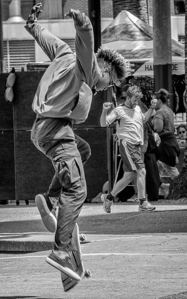 Man jumps in the air from skateboard with his arms folded back like wings. Behind him, a less athletic individual walks along pumping his arms.