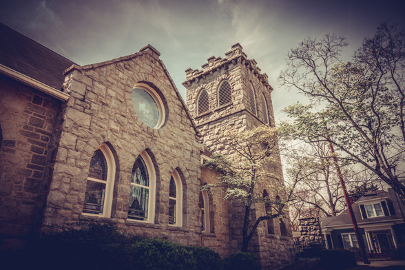 Historic stone church photographed from a low angle against a cloudy sky. The building features rough-cut stone masonry with Gothic Revival architectural elements including three tall arched windows with white frames, a circular rose window in the gable, and a square bell tower with louvered openings and crenellated top. The structure shows weathered stonework in shades of tan and gray. Bare deciduous trees frame the right side of the composition, with a glimpse of a residential building visible in the background. The image has a vintage, sepia-toned quality with dramatic lighting emphasizing the building's texture and verticality.